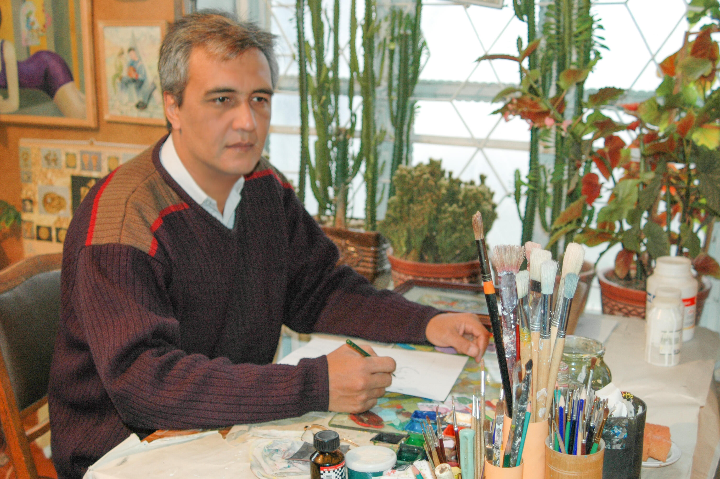 Photo of Olim Kamolzoda sitting at the desk in his workshop. He is the Honoured Artist of the Republic of Tajikistan, a Member of the Union of Artists of the Republic of Tajikistan, and a Specialist of the Miniature Art. Studying the specifics of the work